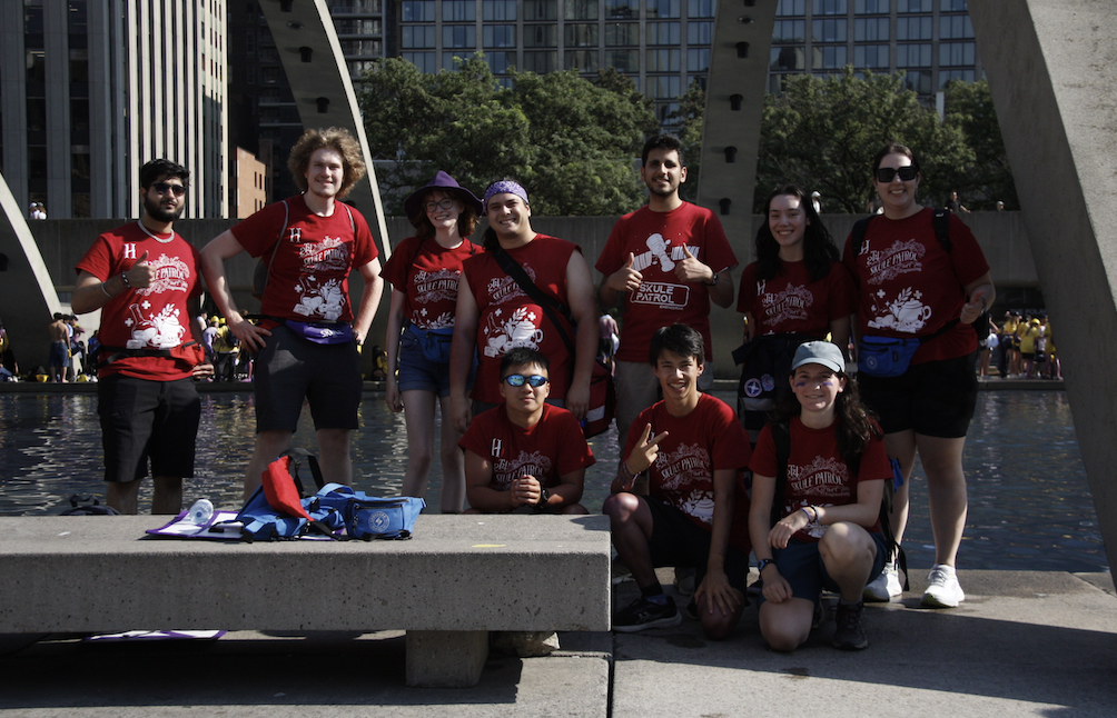 A photo of ten people; the Head Patroller team and Skule Patrol Co-Directors from Frosh Week 2T4 all stand in front of the fountain at Nathan Phillips Square in Toronto after the Downtown Walkaround event. They are all wearing red Skule Patrol shirts and smiling, some give thumbs up or peace signs. Additional Skule Patrol kits sit on the concrete bench in the front of the photo.