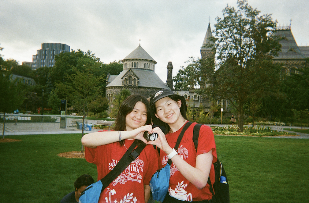 Screenshot 2026-06-03 at 16.47.24 Two Skule Patrollers from 2T4 with blue kits and red Skule Patrol shirts make a heart wiht their hands. They stand on Front Campus with University College behind them,